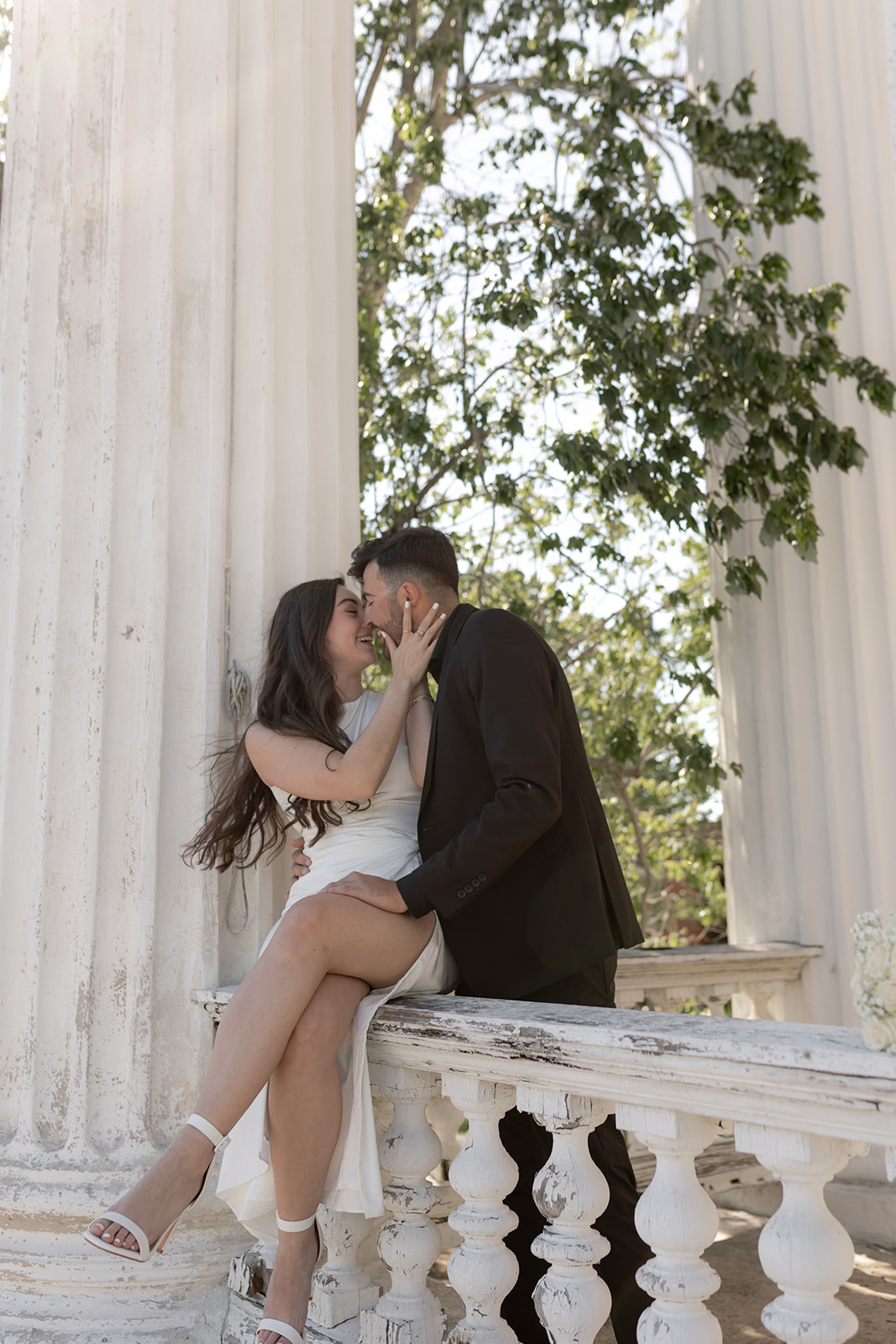 couple in love. Cool black and white engagement attire. Woman is sitting on stone reiling. Man kissing her from behind.