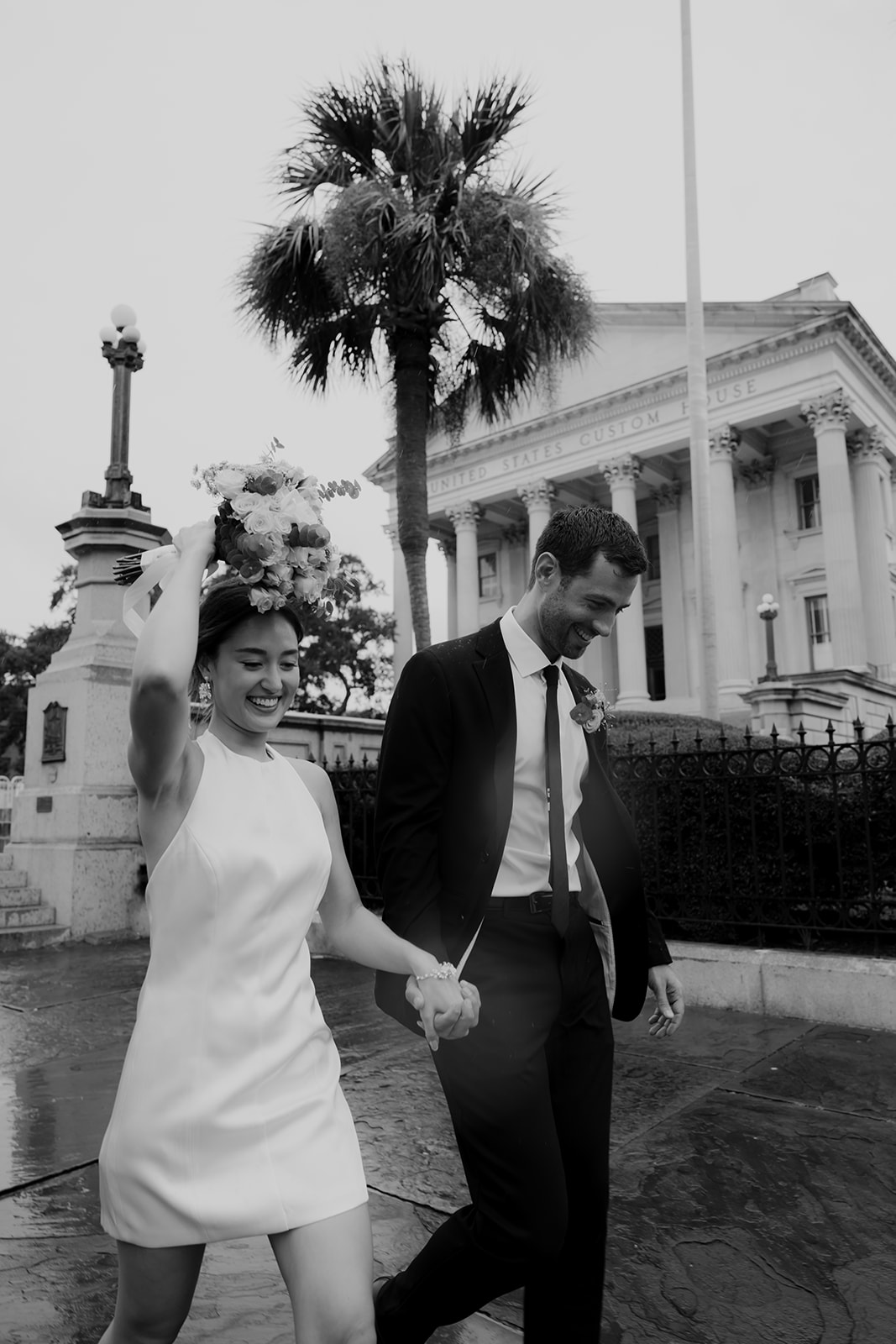 Woman in white neckholder dress is holding flowers over hher head to cover from rain. Man in black suit is walking next to her and holding her hand. US Custom buidling and palmetto tree in background. Stunning Engagement attire Look