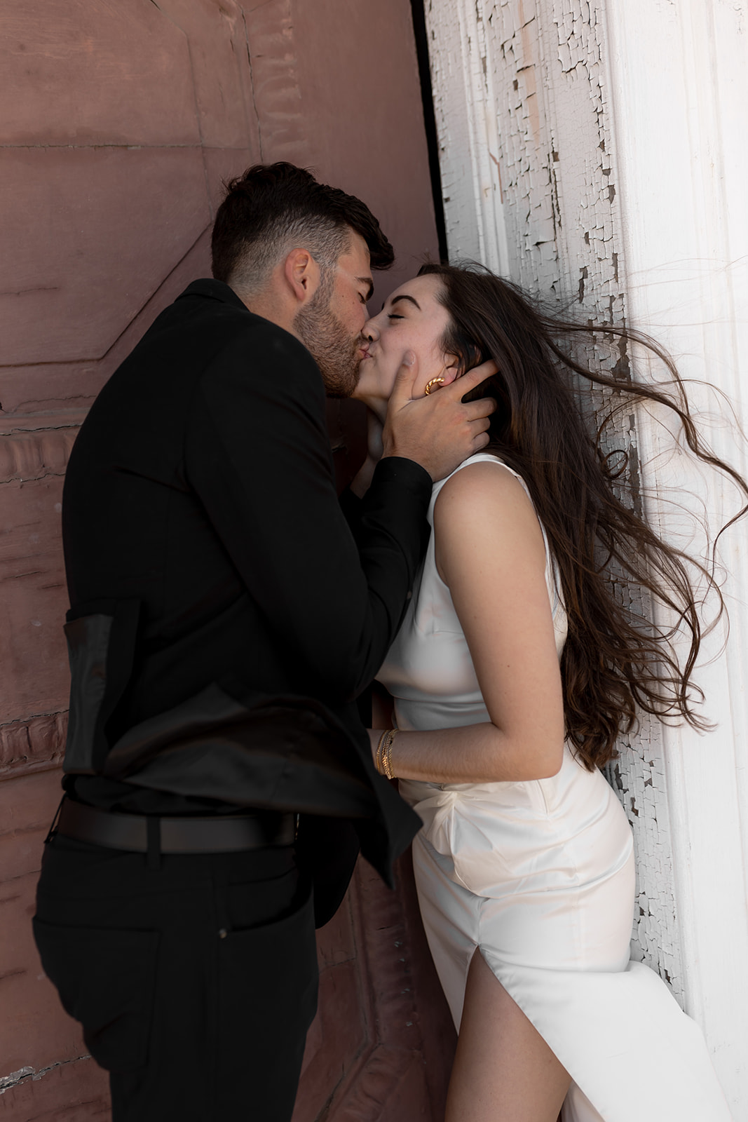 Couple kissing while wind is blowing the womans hair to the side. Man holding her cheeks with both hands. dressed in black and white engagement attire