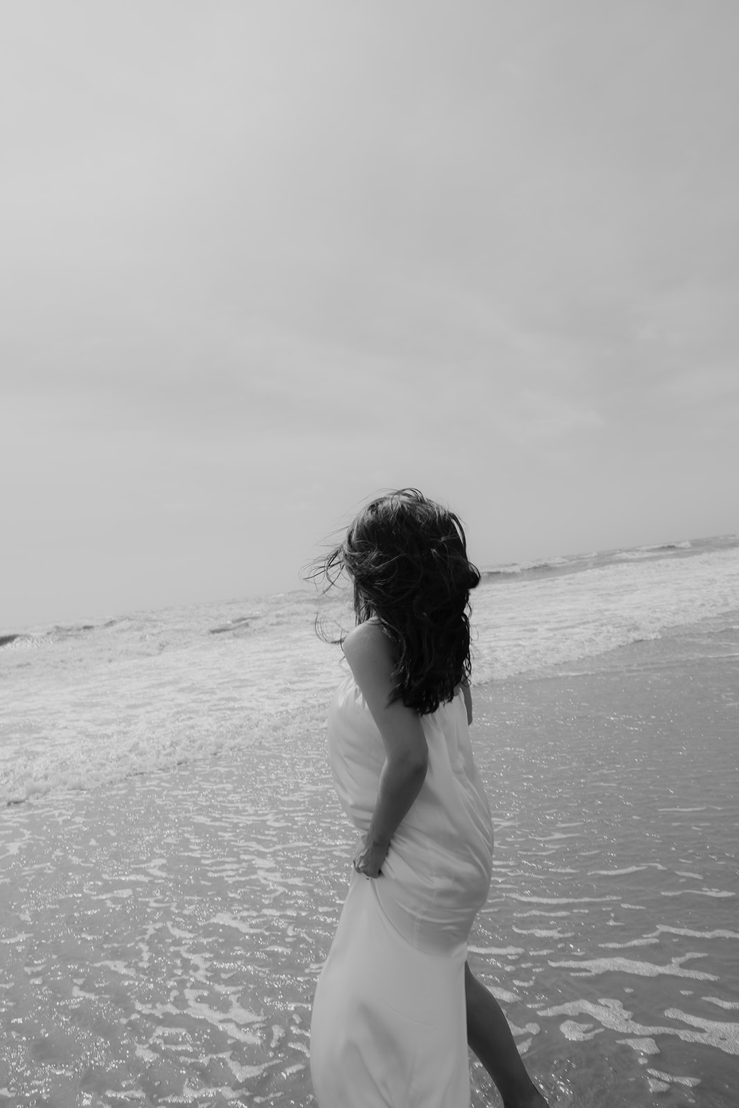 Black and white photo of woman standing at the beach with ocean in front of her. Authentic engagement photos