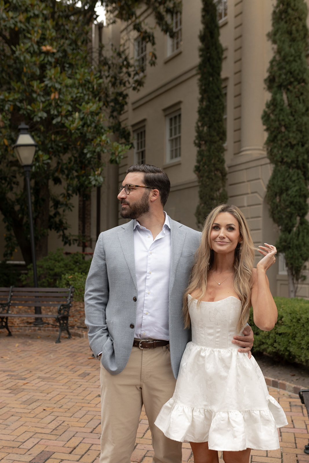 Woman in white dress looking into camera and plays with hair. Man in blue jacket and beige pents looks to the side. Italian cypress in background. Photo captured in Downtown Engagement Charleston