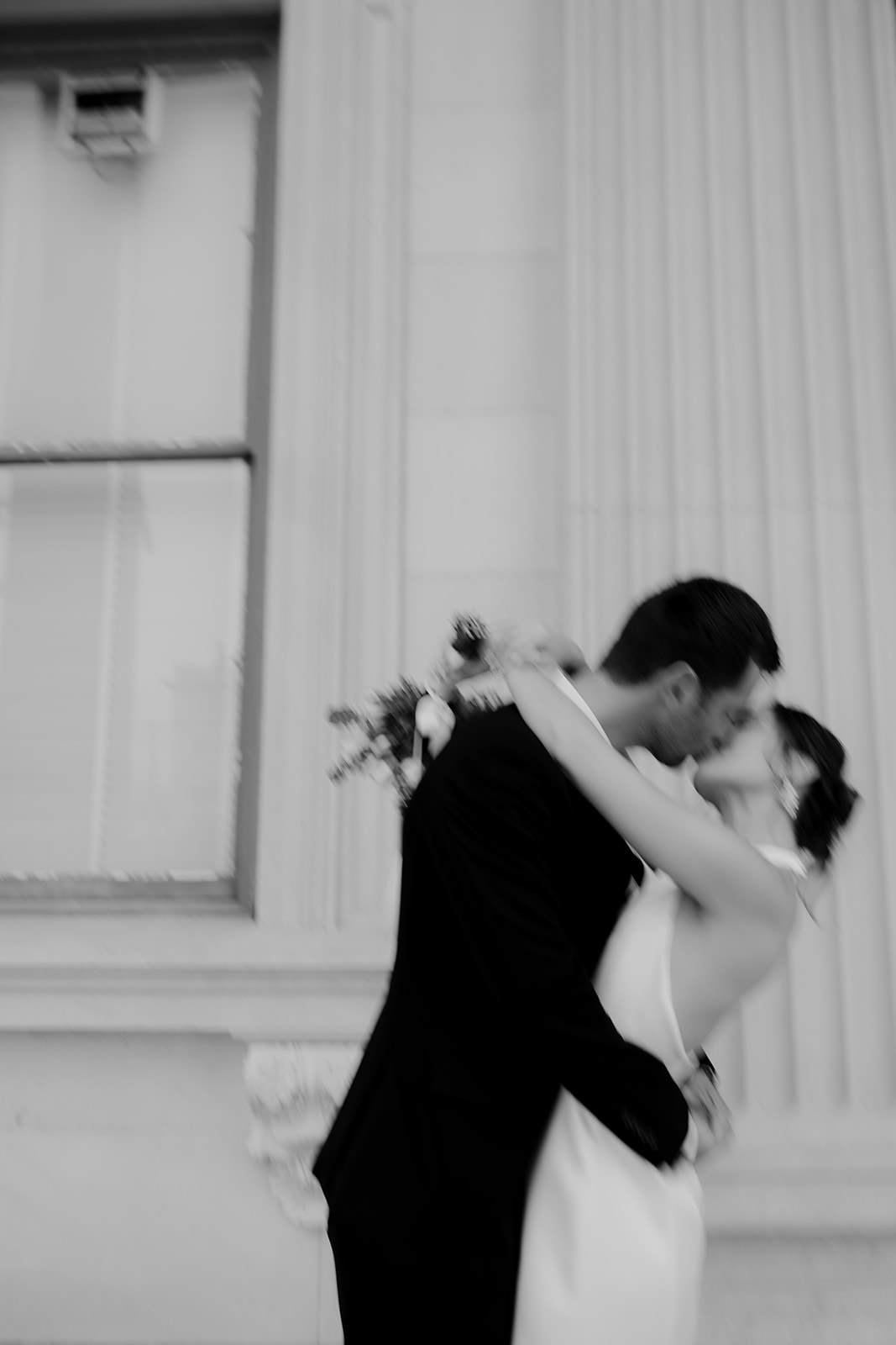 Artsy and blurry photo of couple kissing. Man is slightly dipping the woman which is holding flowers in one hand.