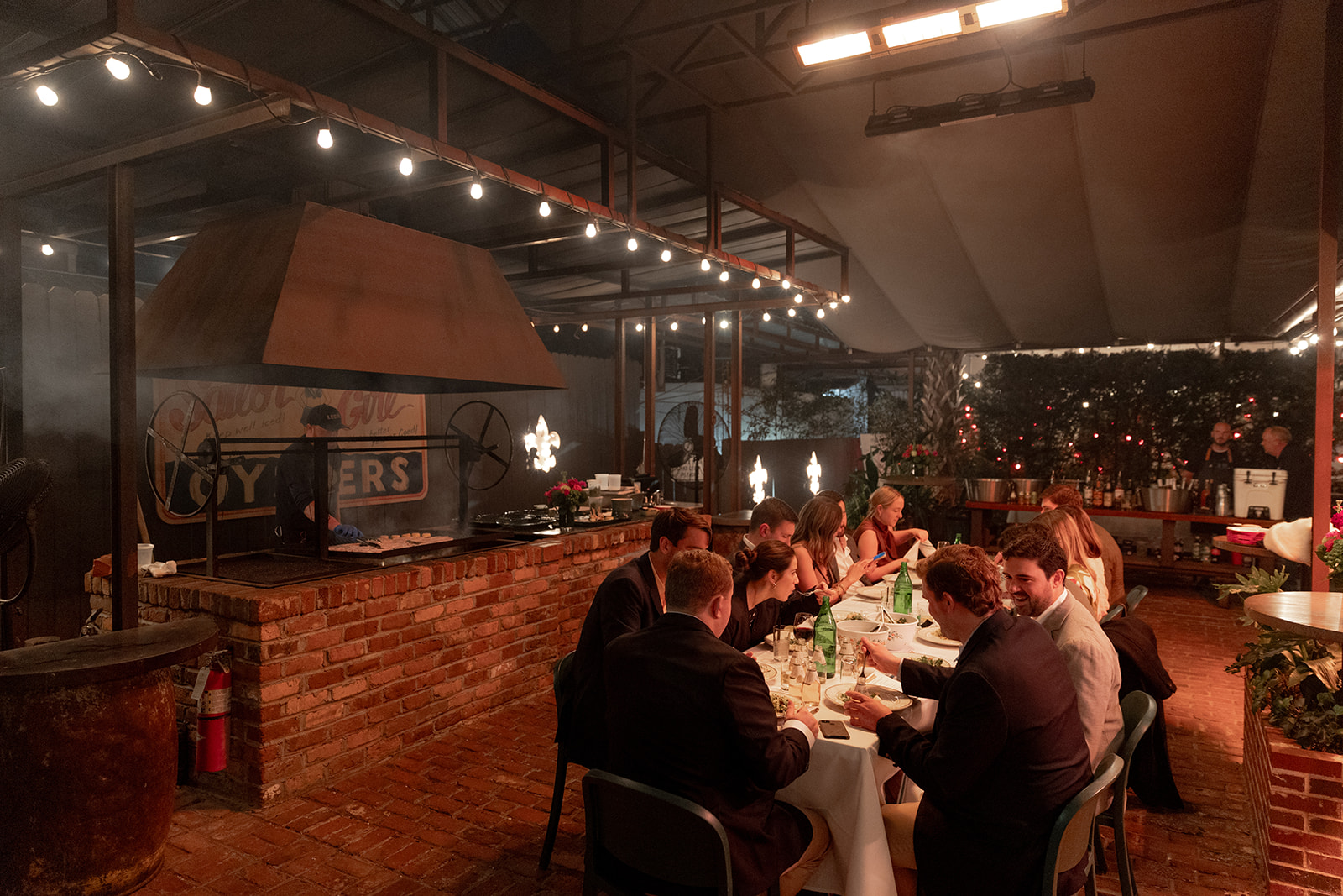 Family style Rehearsal Dinner at Leon's Oyster Shed. Wedding guest sitting in front of open fire grill at long table.