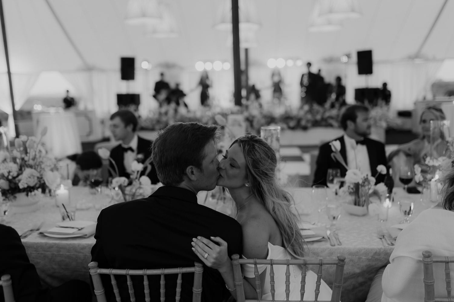 Bride and groom share a kiss sitting at wedding reception captured in black and white
