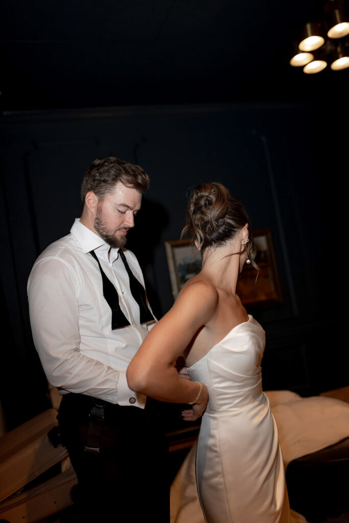 The groom with open bowtie is helping the bride opening the dress after reception before switching into her second dress