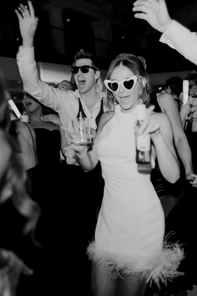 Bride with heart glasses and a dink in her hand is smiling towards the camera